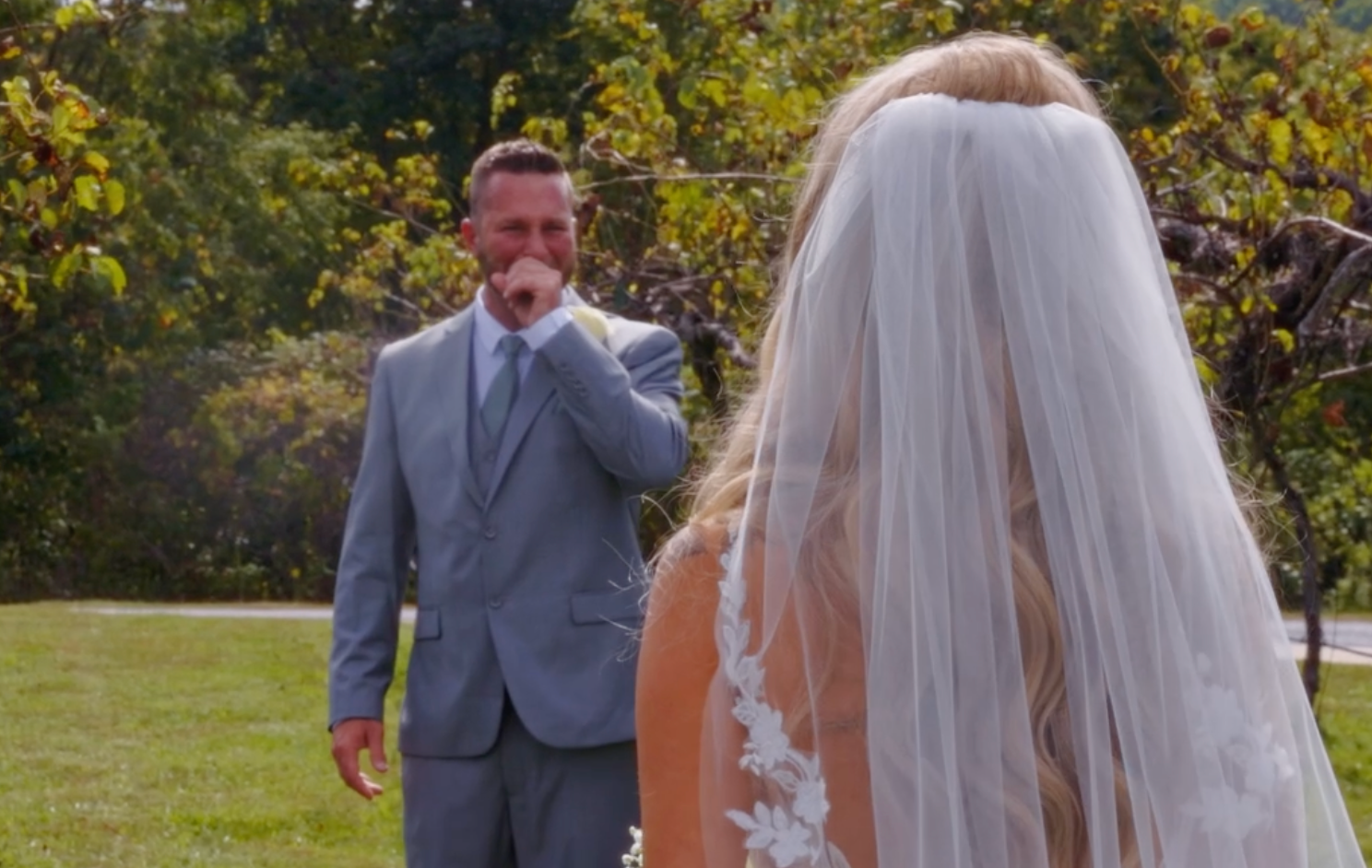 Emotional groom in gray suit reacts with tears as bride in white lace gown and veil approaches during outdoor wedding ceremony.