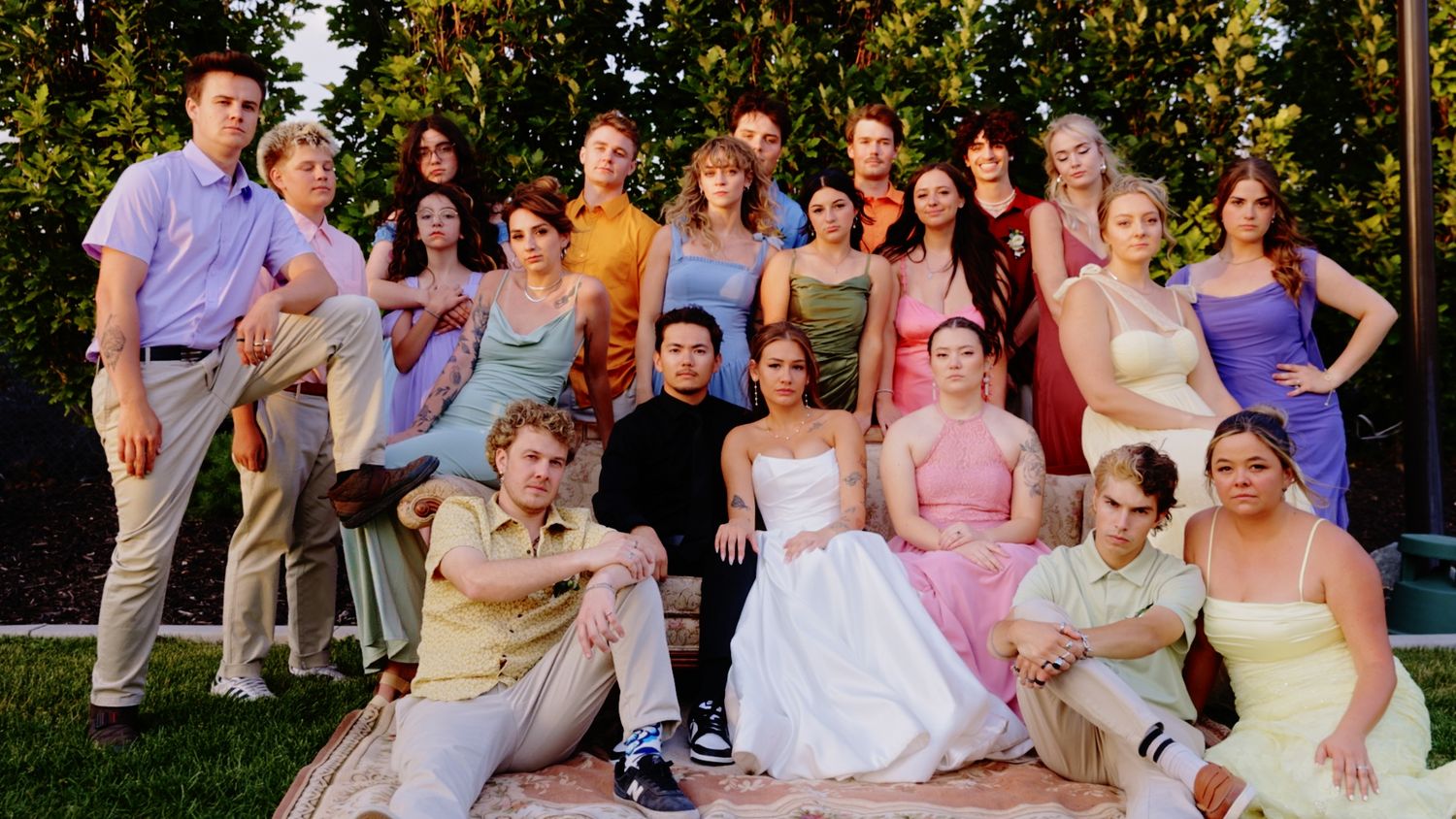 Large wedding party group photo outdoors, bride in white gown surrounded by guests in colorful formal attire under trees.