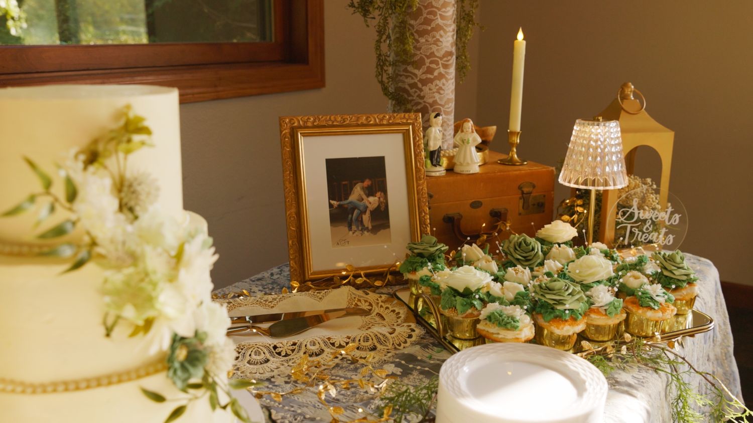 Elegant wedding dessert table with tiered white cake, cupcakes, candles, and a gold-framed photo display.