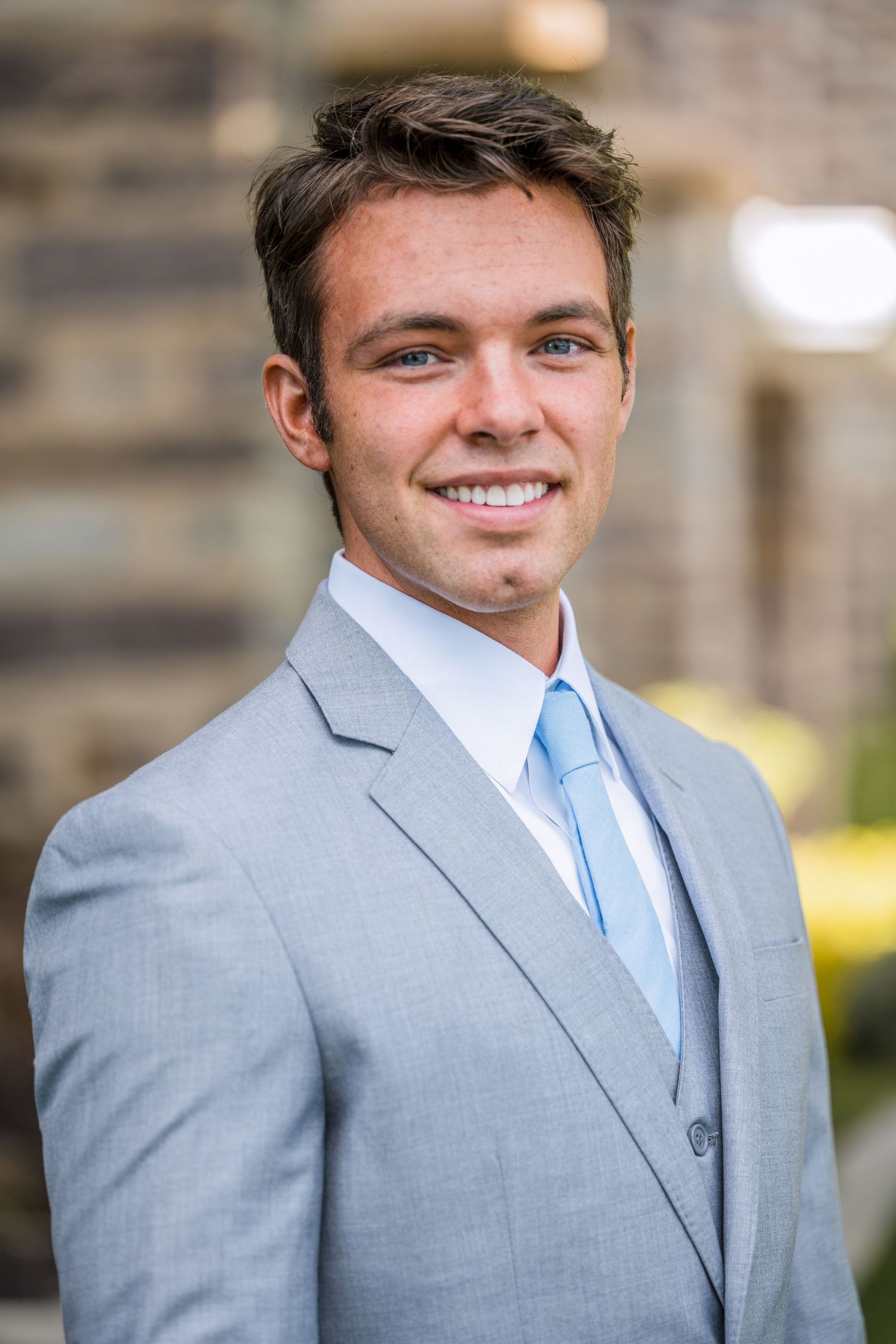 Professional headshot of someone in a light gray suit and baby blue tie smiling at the camera outdoors.
