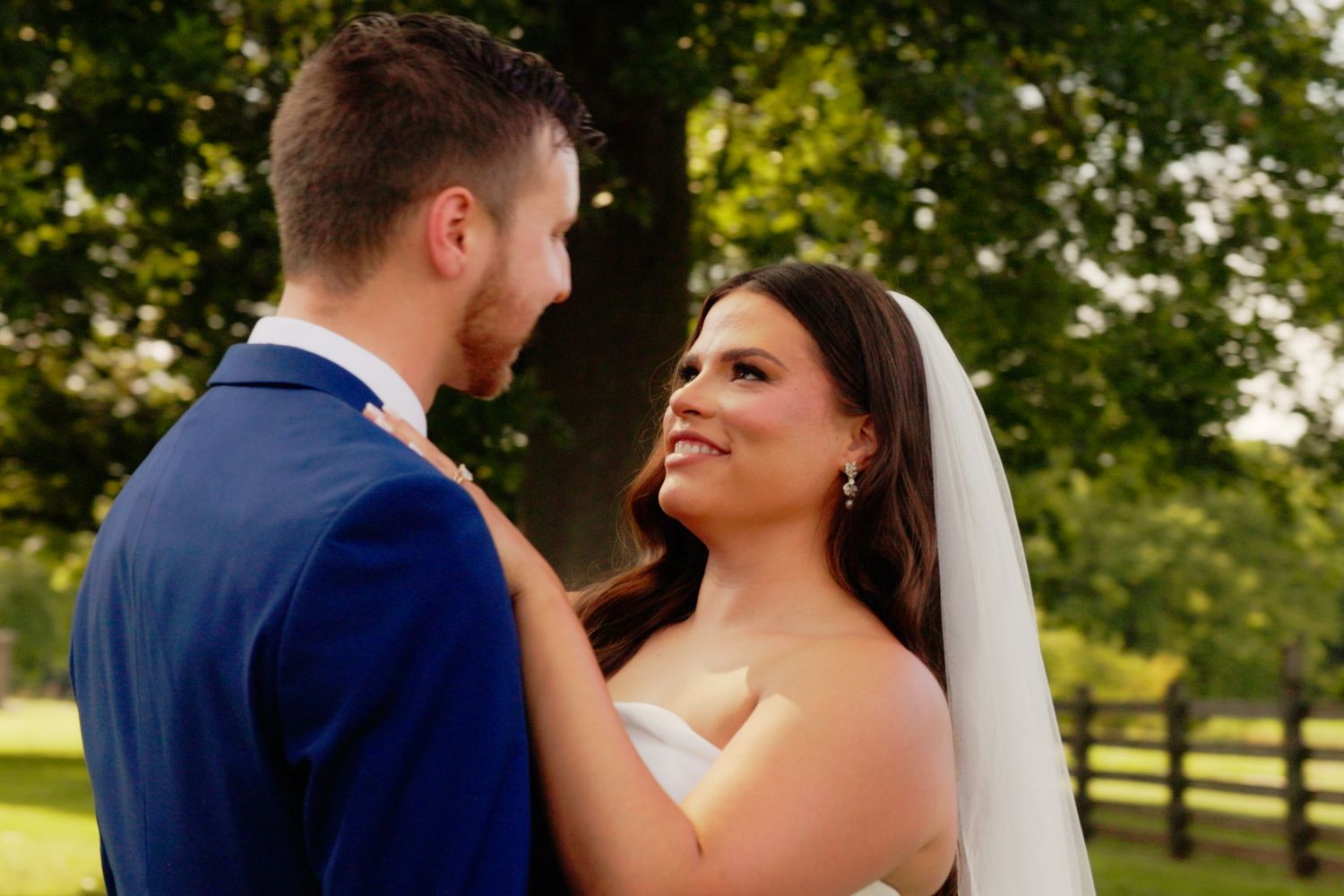 Bride in white veil smiles at groom in blue suit during outdoor wedding ceremony surrounded by greenery.
