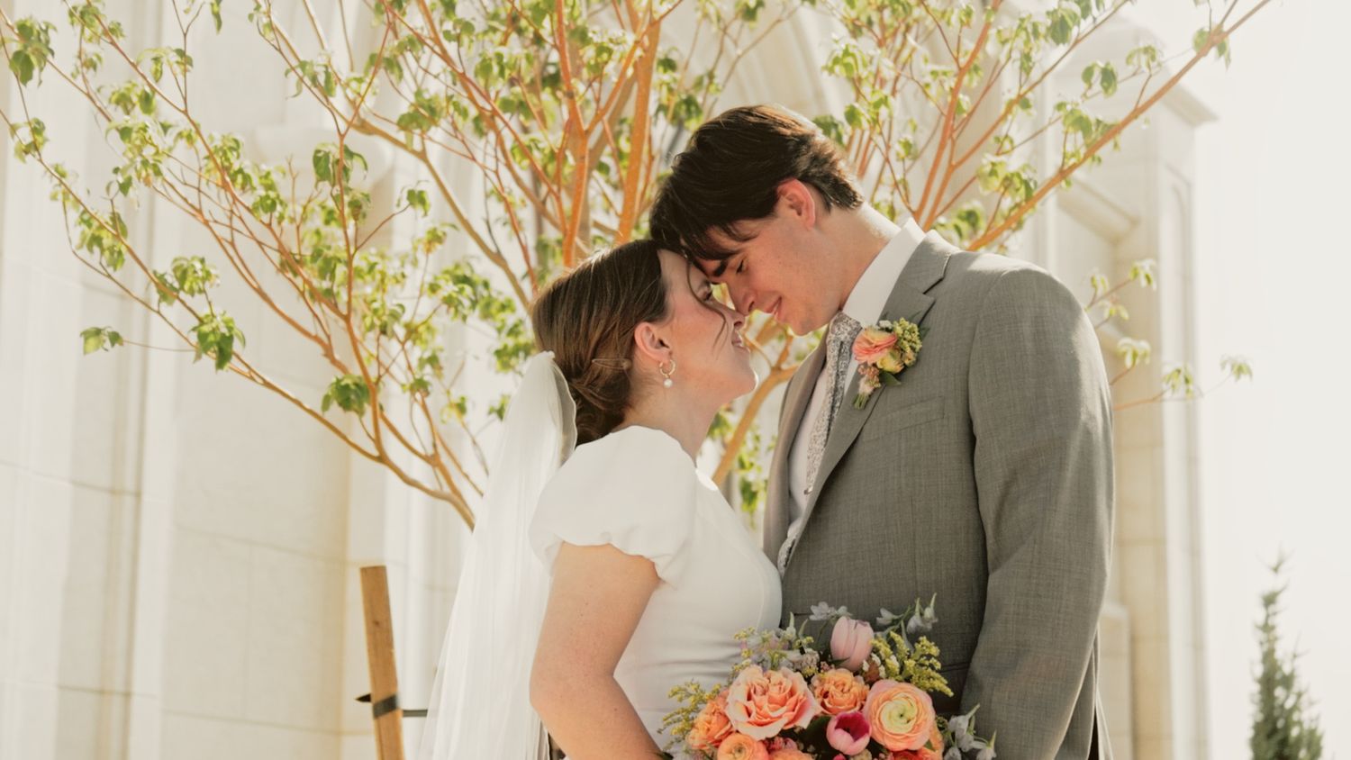 A romantic wedding moment in front of white walls with spring branches and peach-colored floral bouquet.