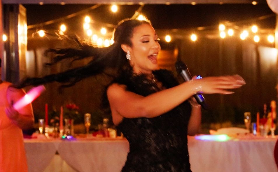 Woman in black dress singing passionately into microphone at elegant event with string lights.