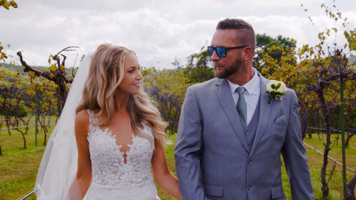 A bride and groom walk together in a vineyard, she in a lace gown and veil, he in a gray suit with sunglasses.