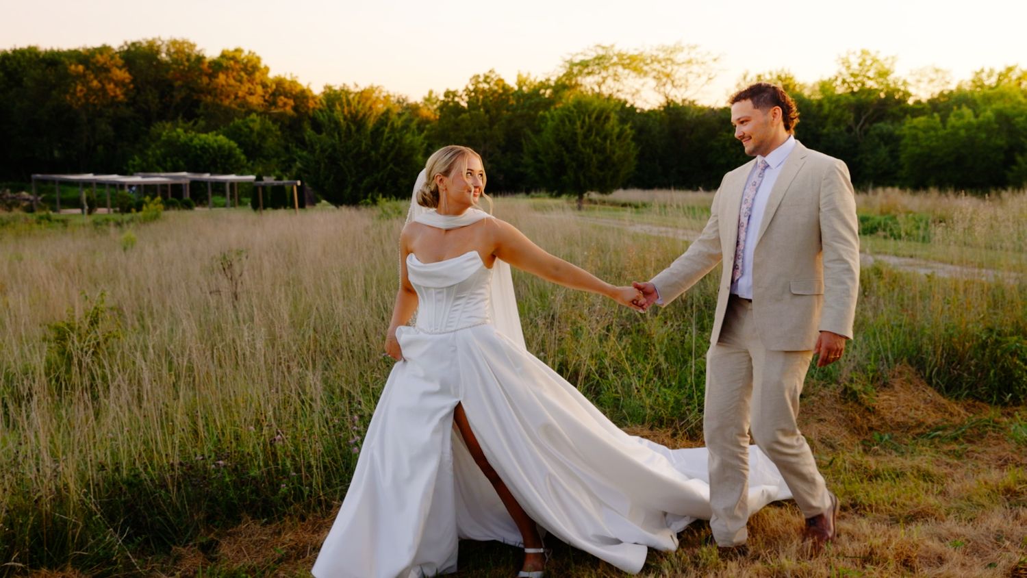 Bride and groom holding hands in golden field at sunset, bride in white gown, groom in tan suit.