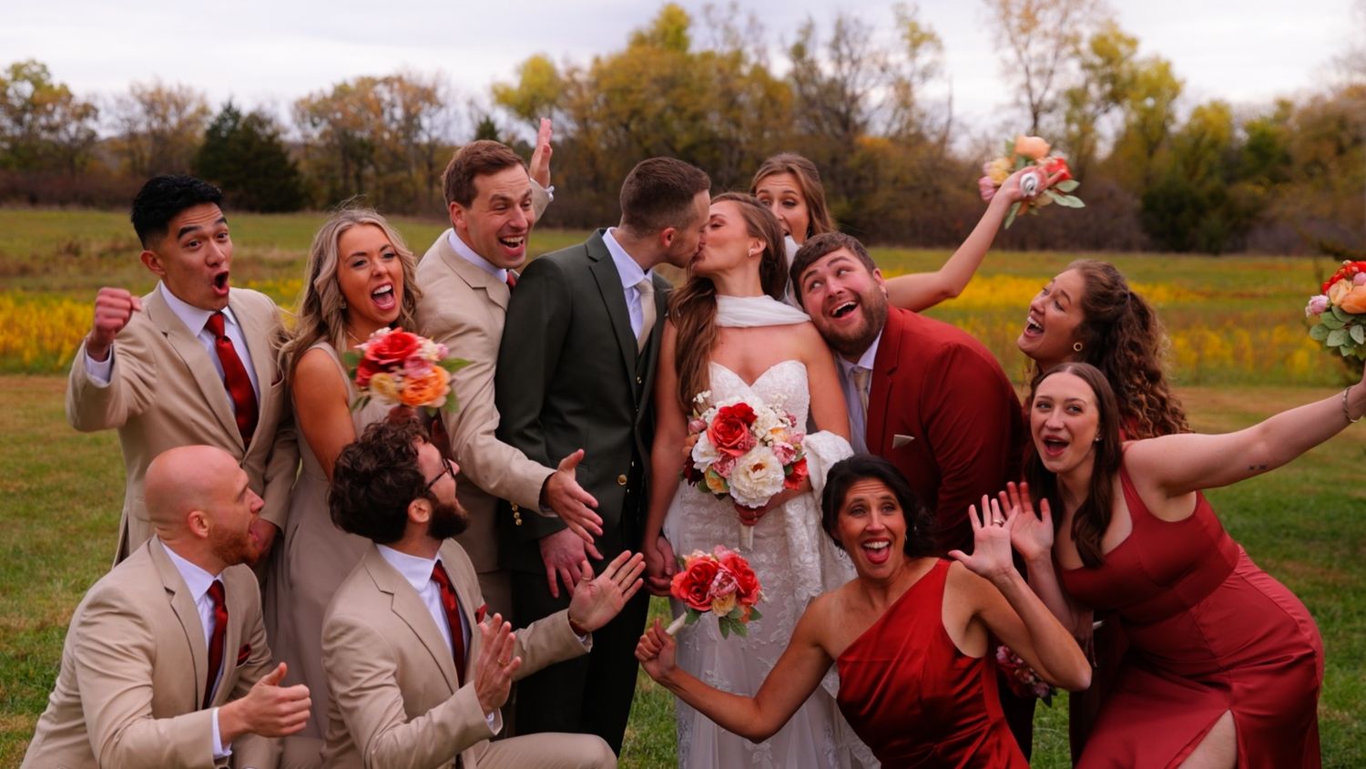 Joyful wedding party celebrating outdoors in autumn, bride and groom kissing surrounded by guests in red and tan attire.