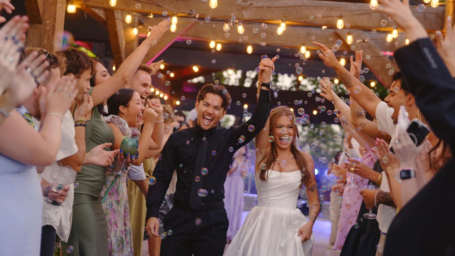 Newlywed couple walks through bubble send-off tunnel at romantic outdoor wedding reception with string lights.