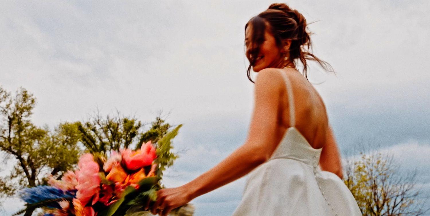 Bride in white backless gown holds colorful bouquet of pink and orange flowers outdoors under cloudy sky.