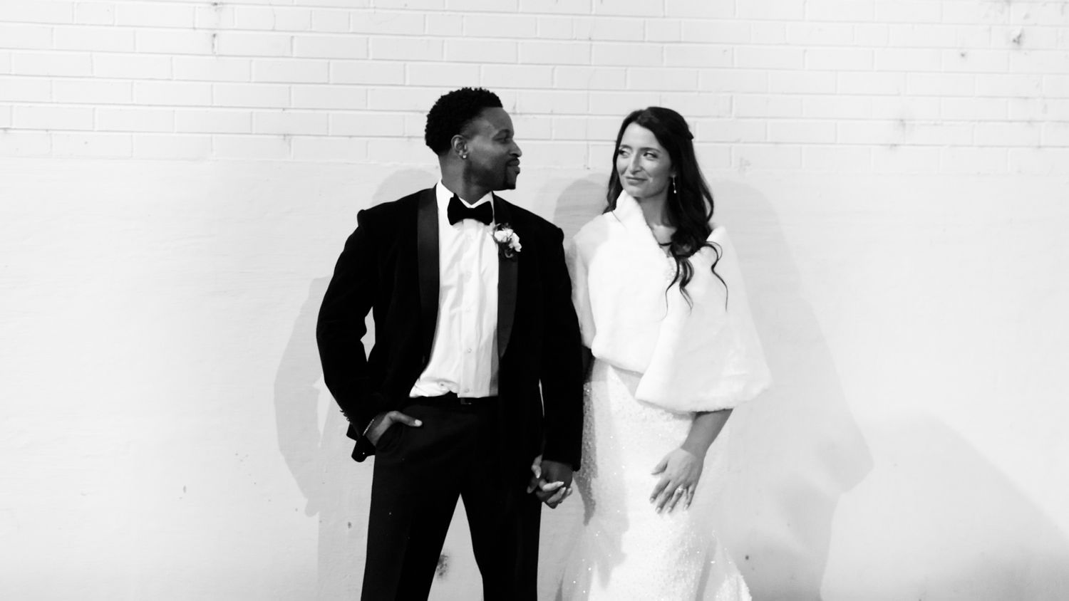 Black and white wedding photo of a couple in formal attire holding hands and smiling at each other.