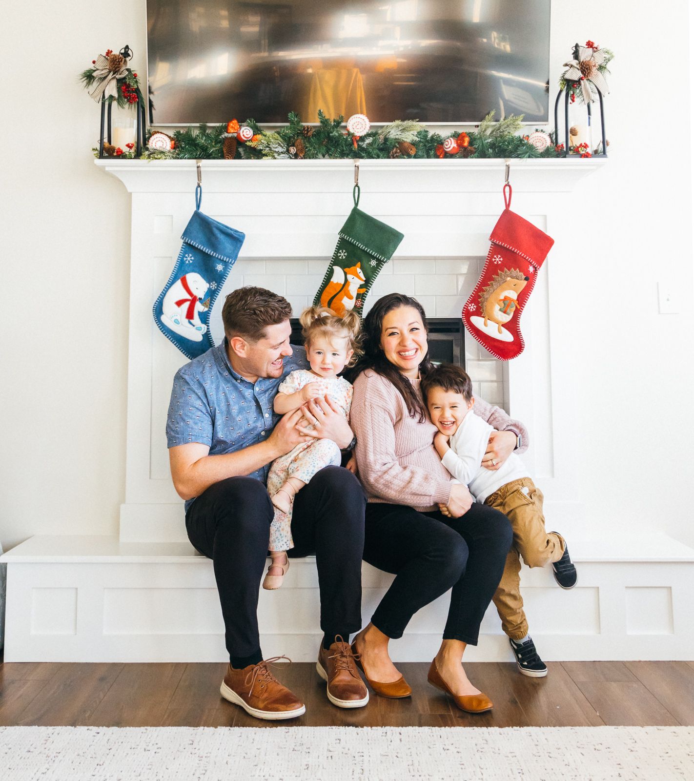 A family sits together by a decorated Christmas fireplace with stockings hung above.