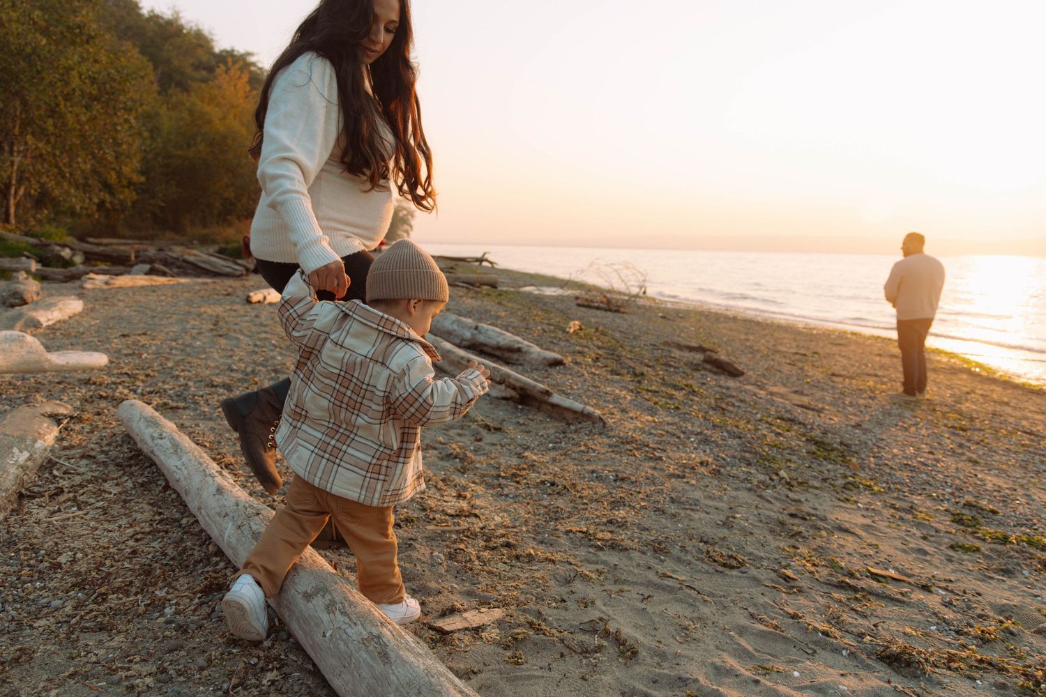 Parent and toddler walking together on driftwood at sunset along a rocky beach.