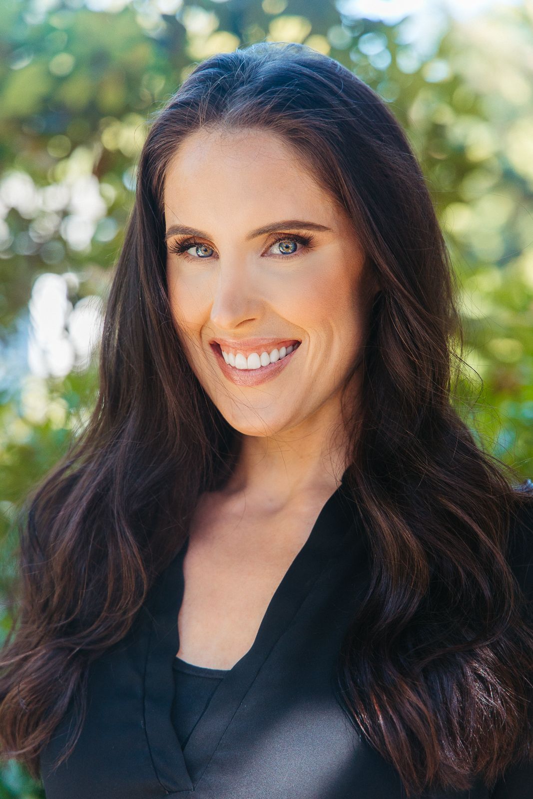 Professional headshot of someone in a black top against a blurred outdoor background.