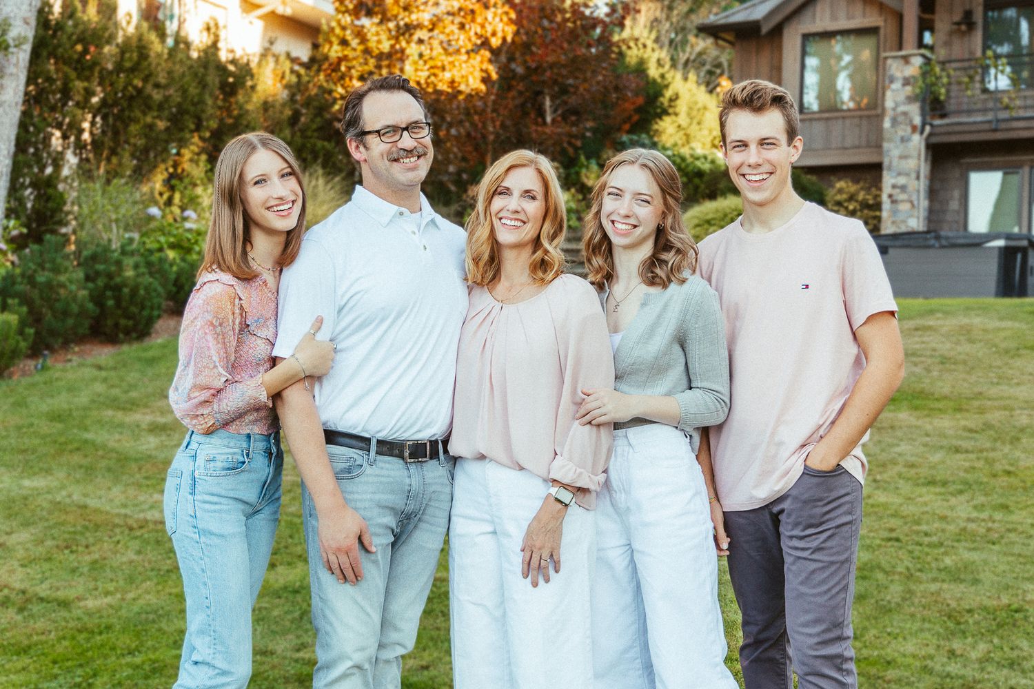 Family group photo with five people in coordinated pastel outfits standing on lawn at sunset.
