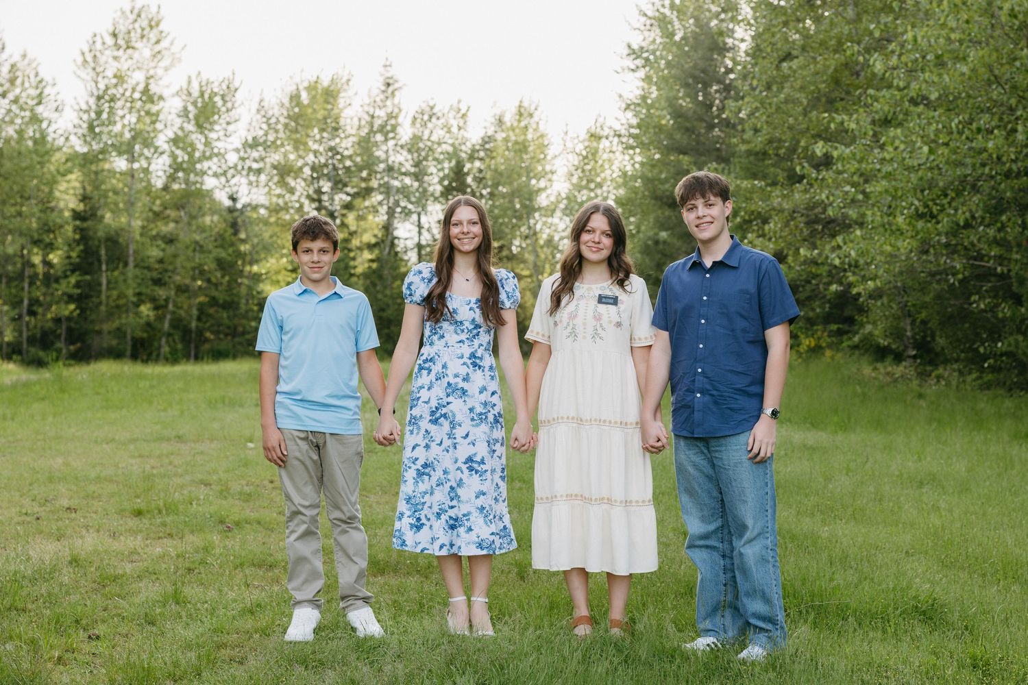 Four people stand in a row on green grass wearing casual spring outfits with trees in the background.