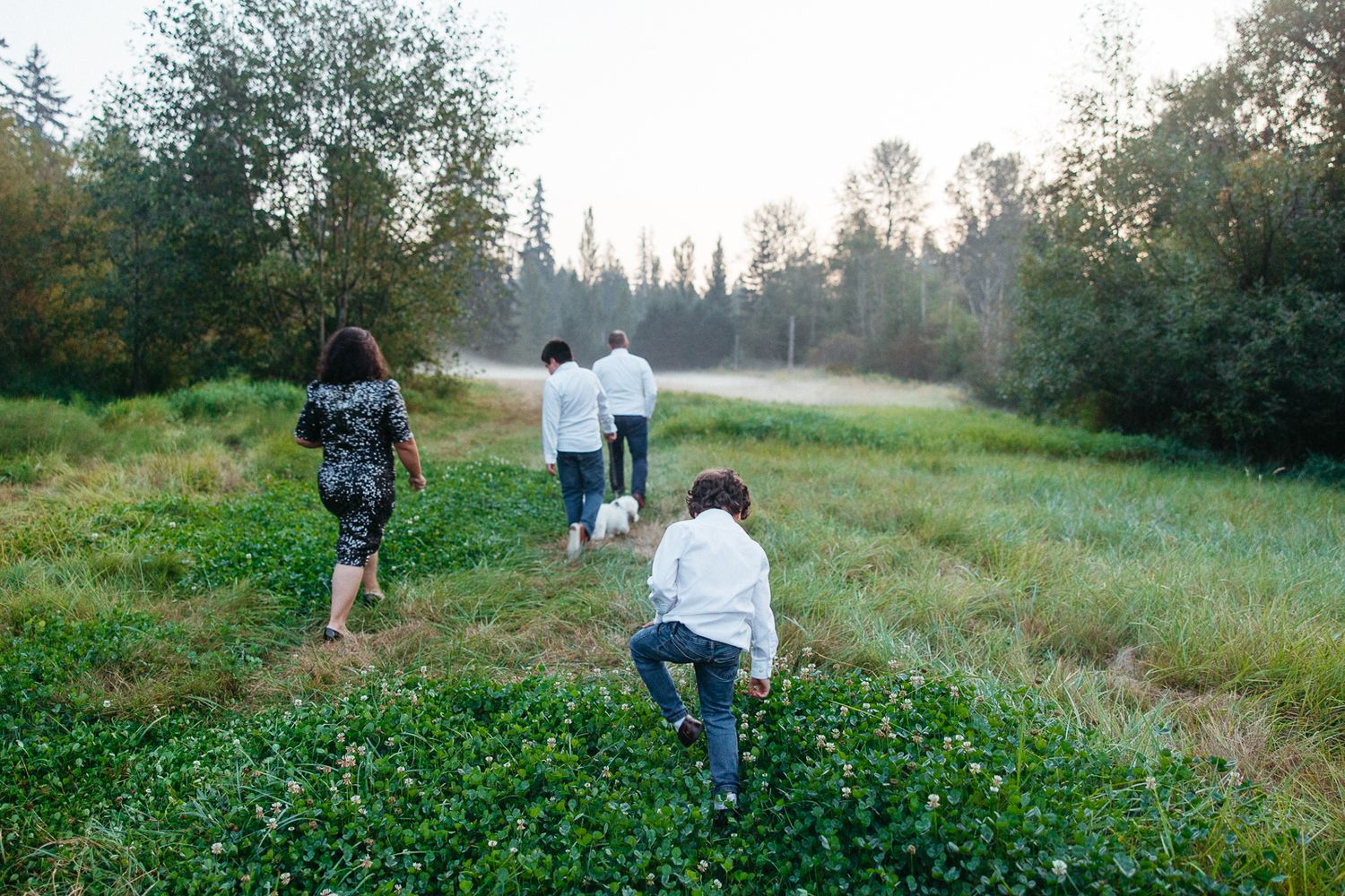 People walking through a lush green meadow with wildflowers as the sun sets behind trees.