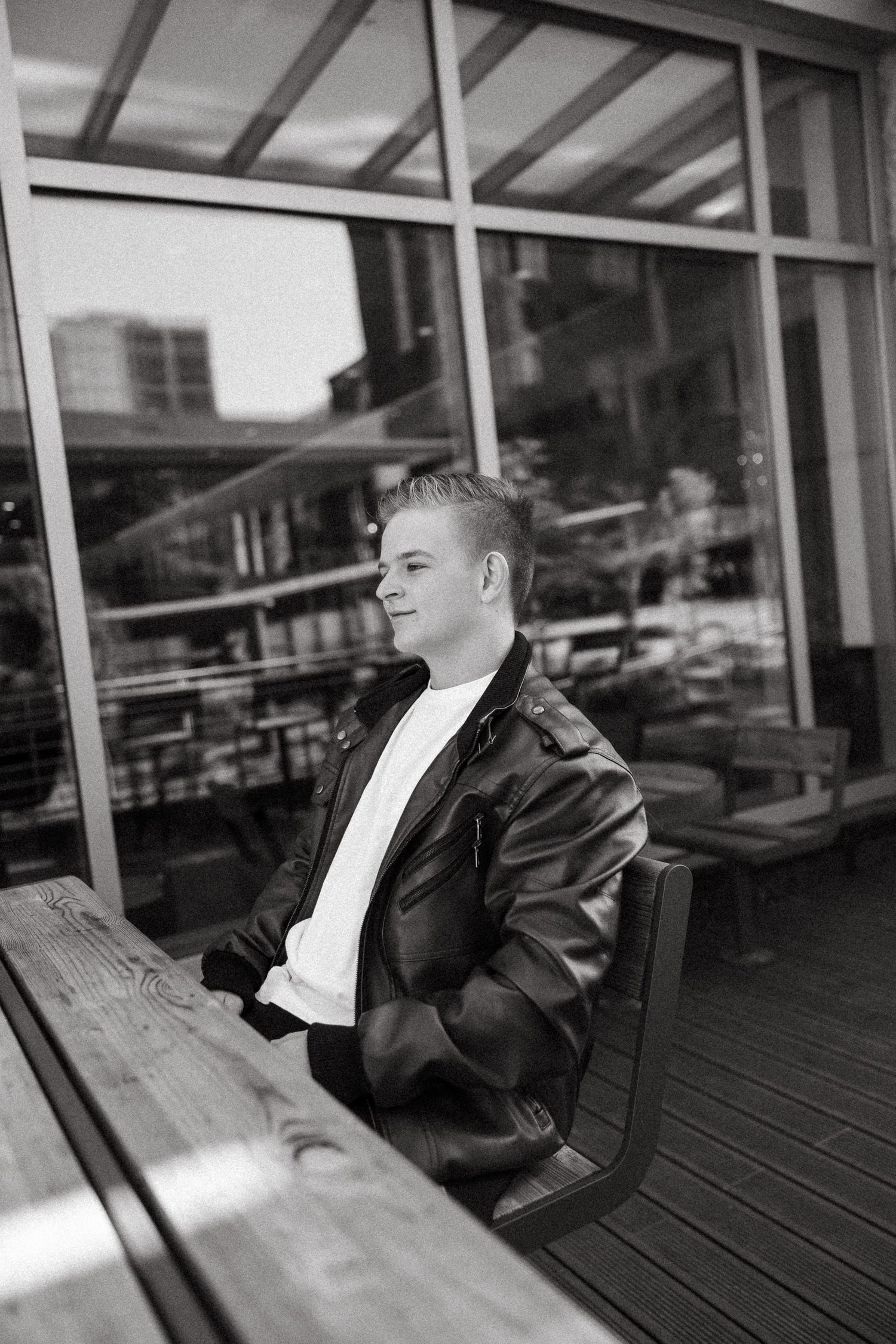 Black and white portrait of someone sitting at a wooden table wearing a leather jacket and white shirt, looking to the side.