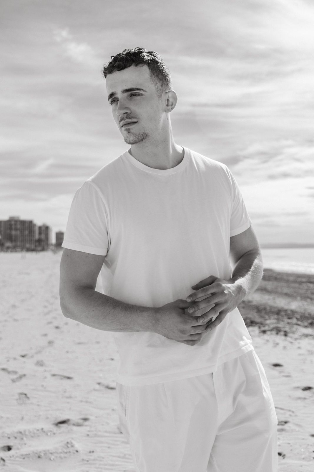 Black and white beach portrait in casual white outfit against cloudy sky.