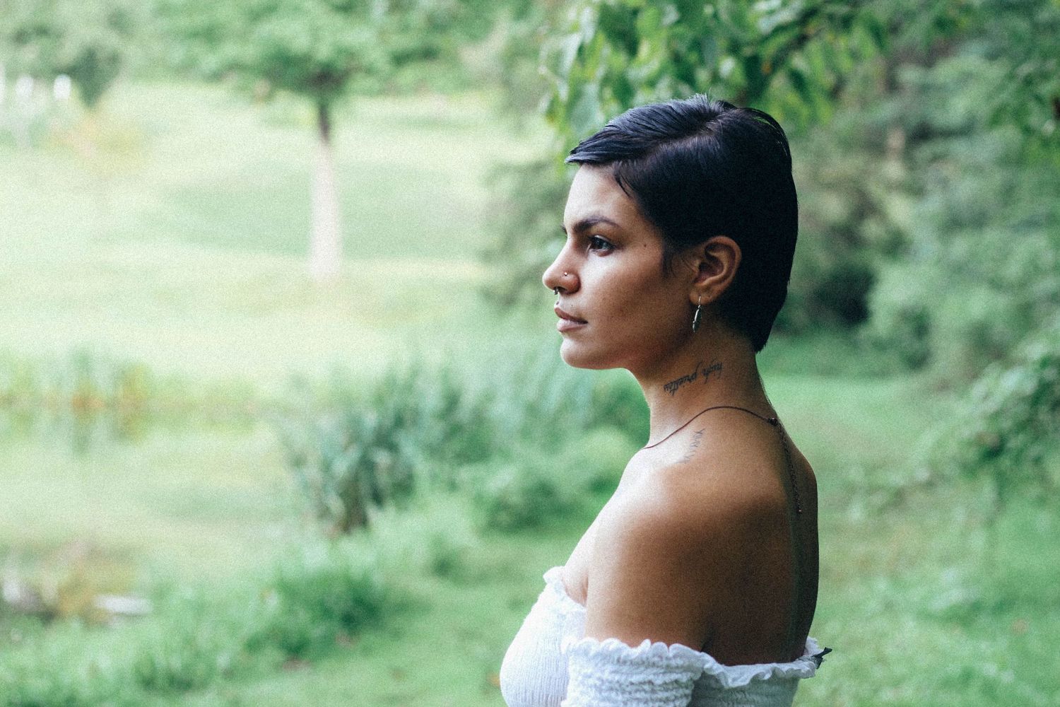 Profile view of person with short dark hair wearing off-the-shoulder white top against blurred green outdoor background.