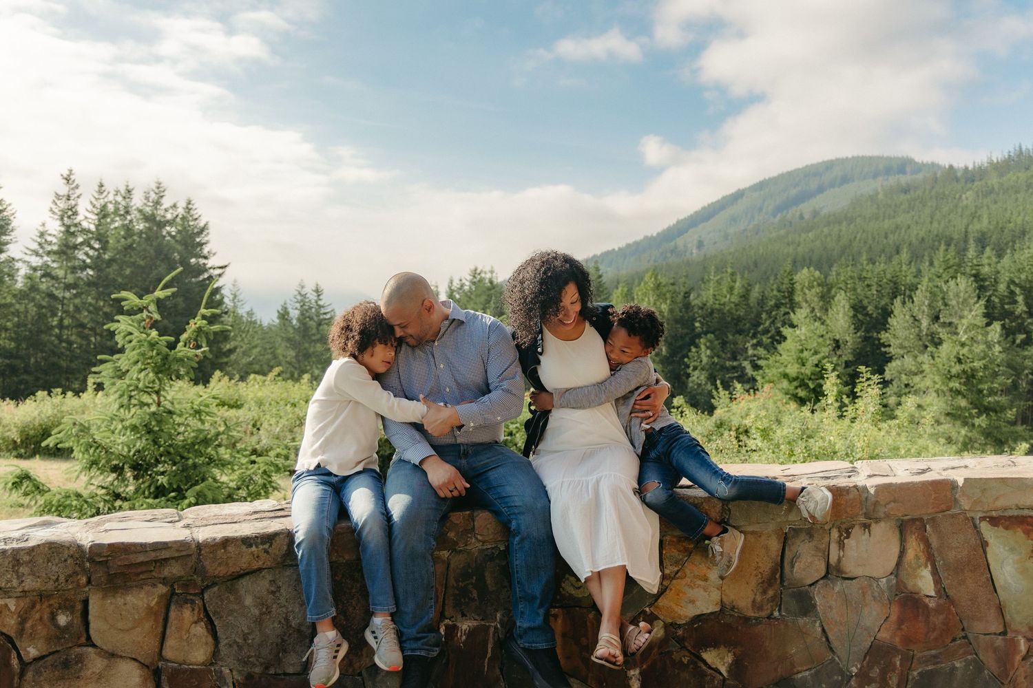 A family sits together on a stone wall overlooking mountains and evergreen forest on a sunny summer day.