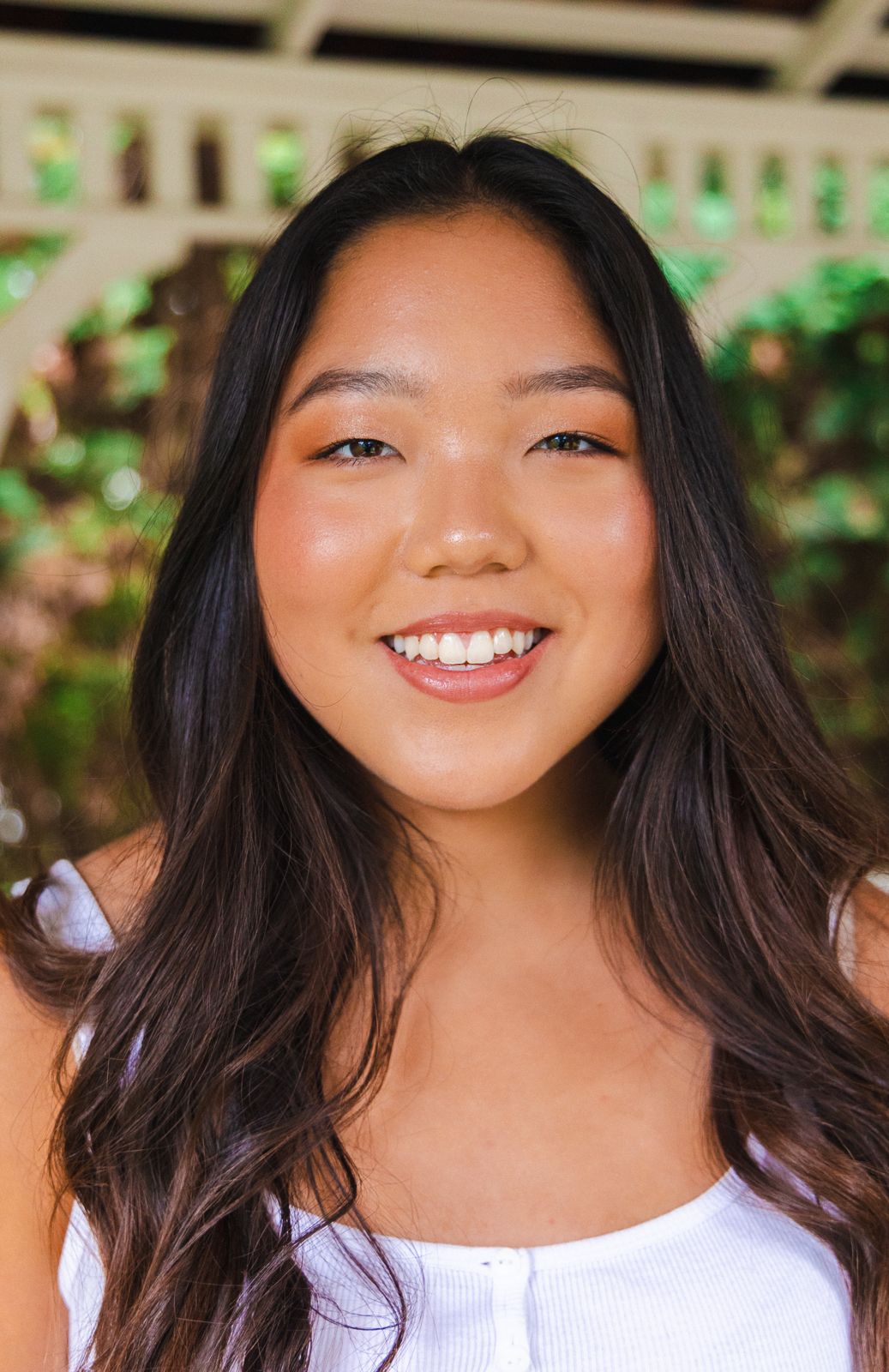 Woman with long dark hair wearing white top smiles brightly outdoors. Makeup services done by Nerissa.