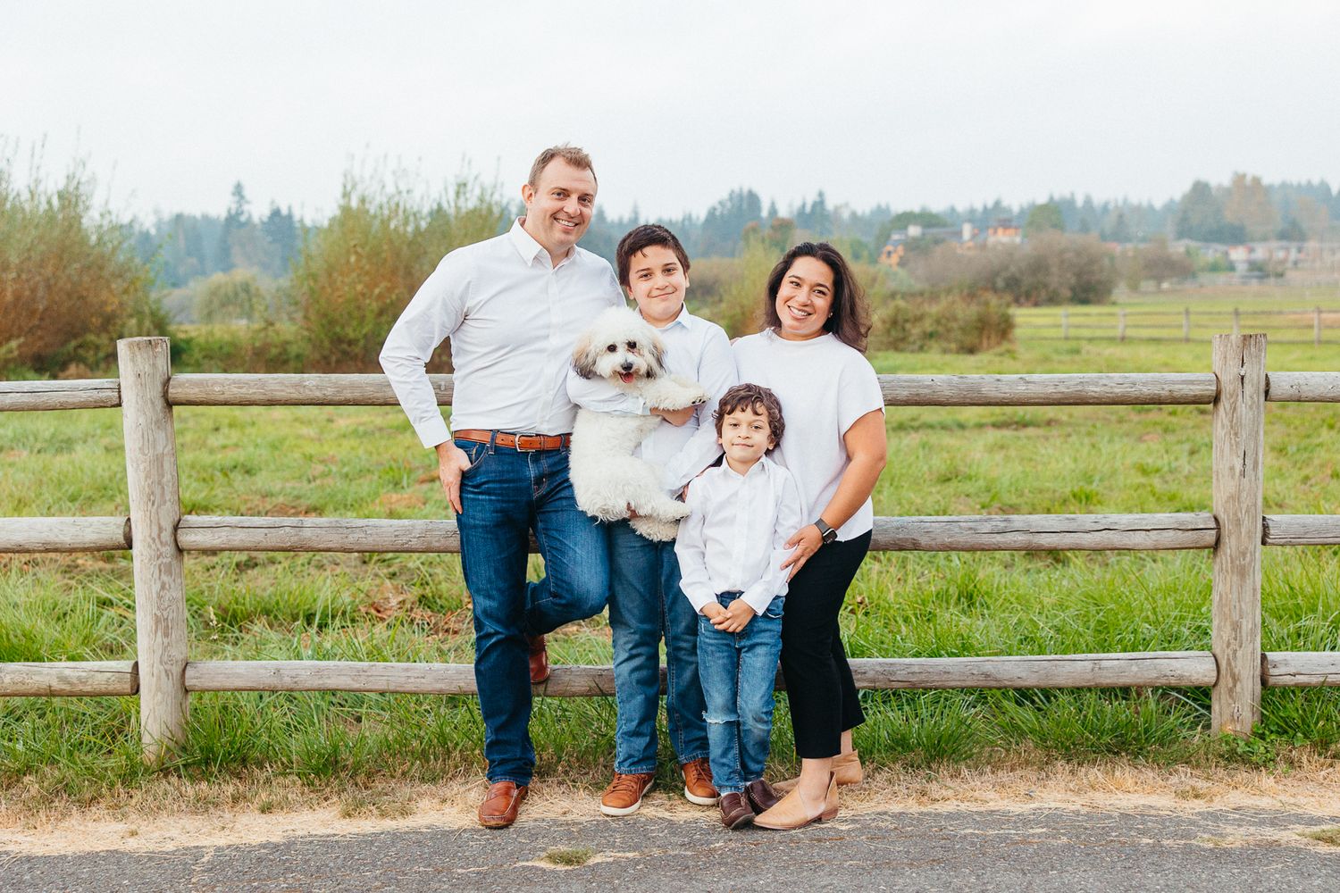 Family of four posing together by a rustic wooden fence in a rural countryside setting.