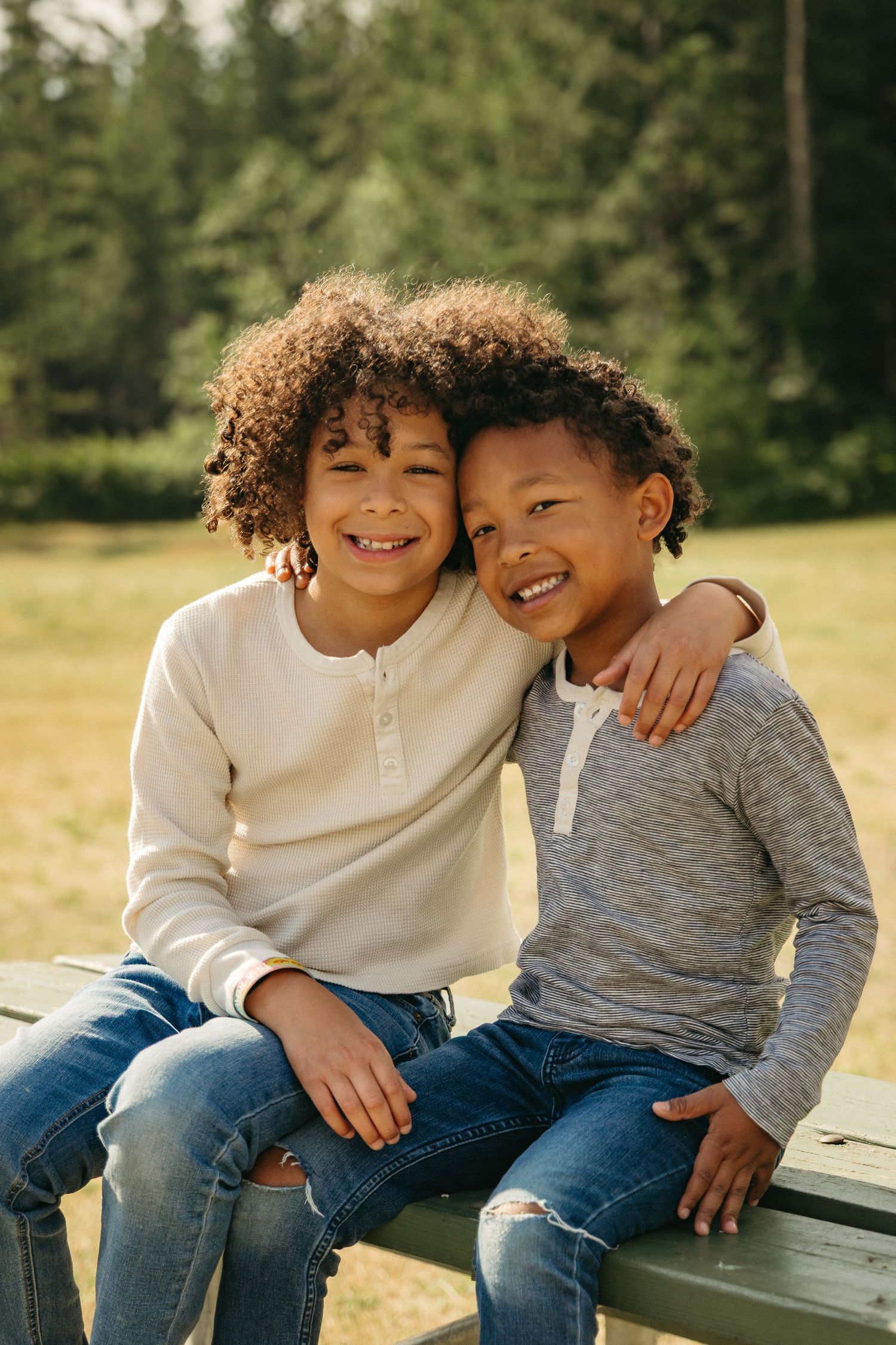Two siblings in casual wear share a playful moment outdoors while sitting together on a sunny day.