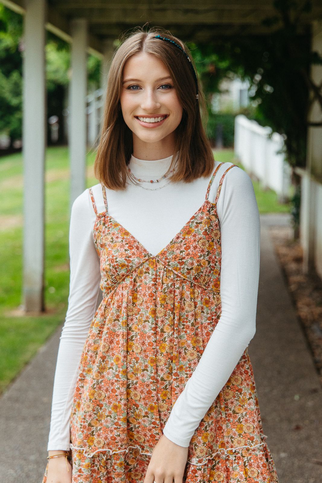 Person wearing white long sleeve top and floral dress standing in covered walkway.