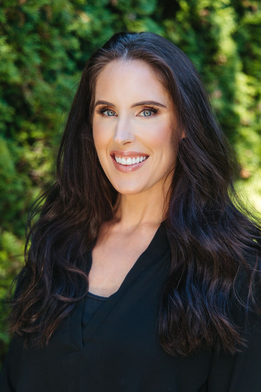 Professional headshot of woman with long dark hair and bright smile against blurred green outdoor background.