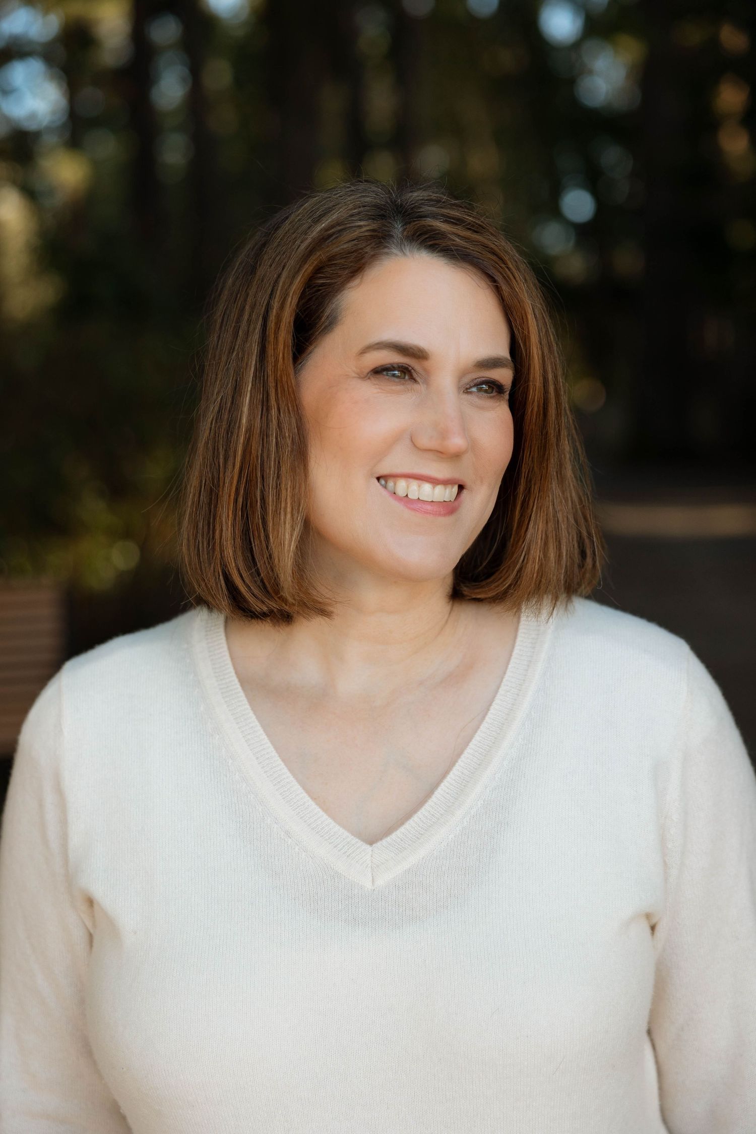 Portrait of woman in white sweater with shoulder-length brown hair smiling outdoors.