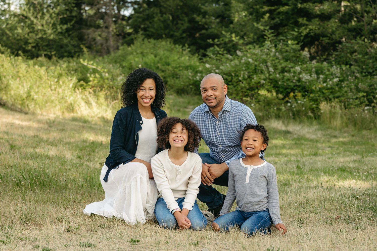 A happy family wearing casual clothing sits together on grass in a sunny outdoor setting during a portrait session.