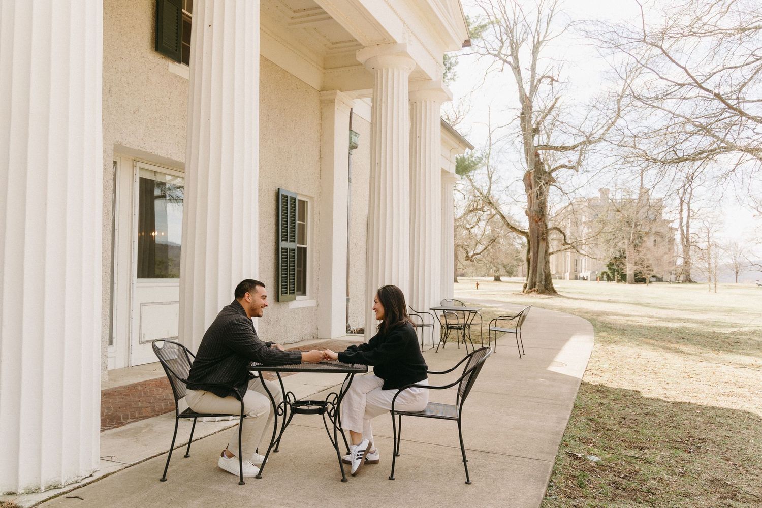 Two people sit at a round table on an elegant porch with white columns in a casual outdoor setting.