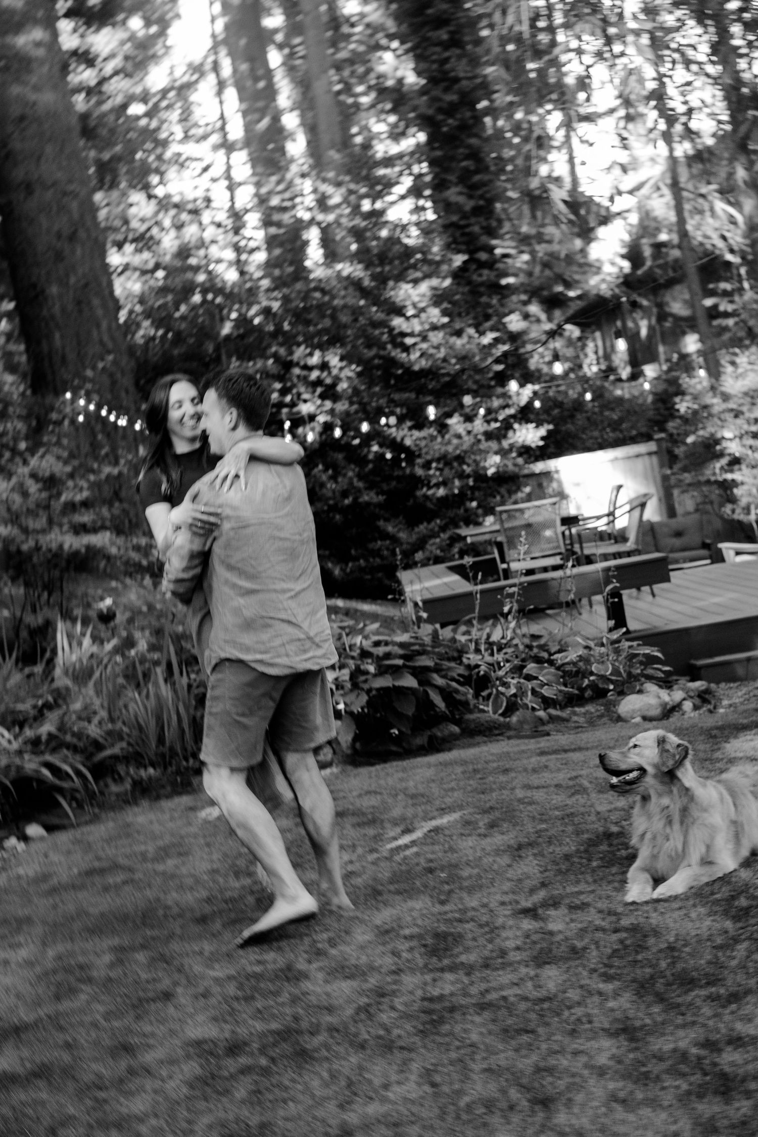 A black and white candid moment in a backyard with string lights and a dog.