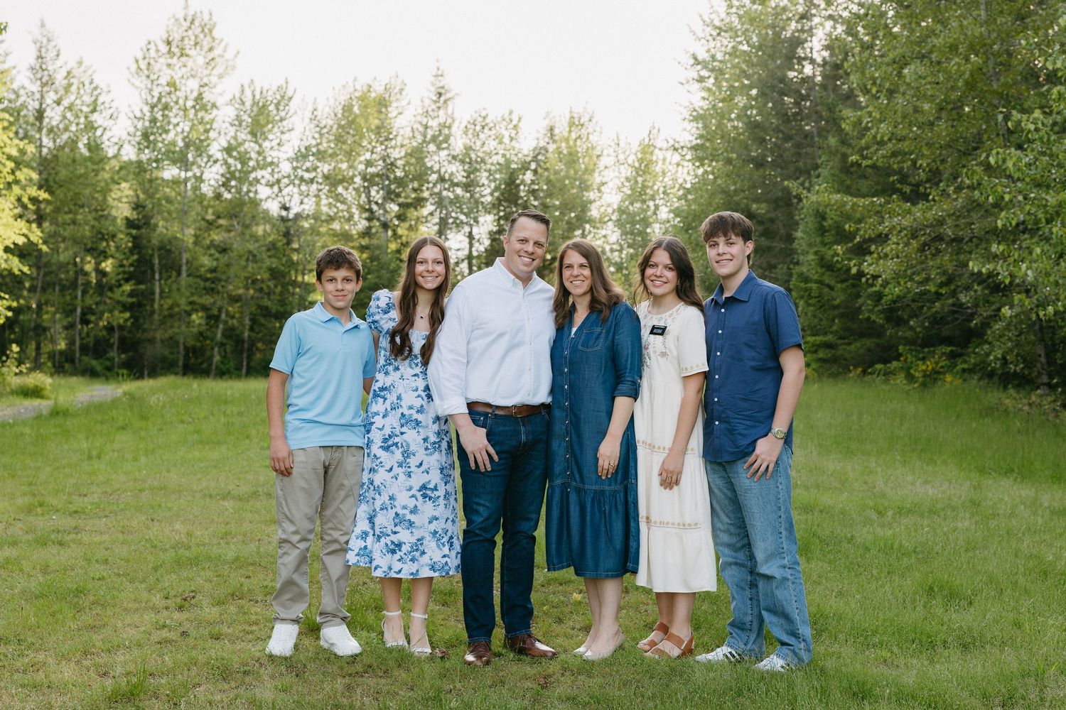 Group gathered on grassy field with evergreen trees in background during summer photoshoot.