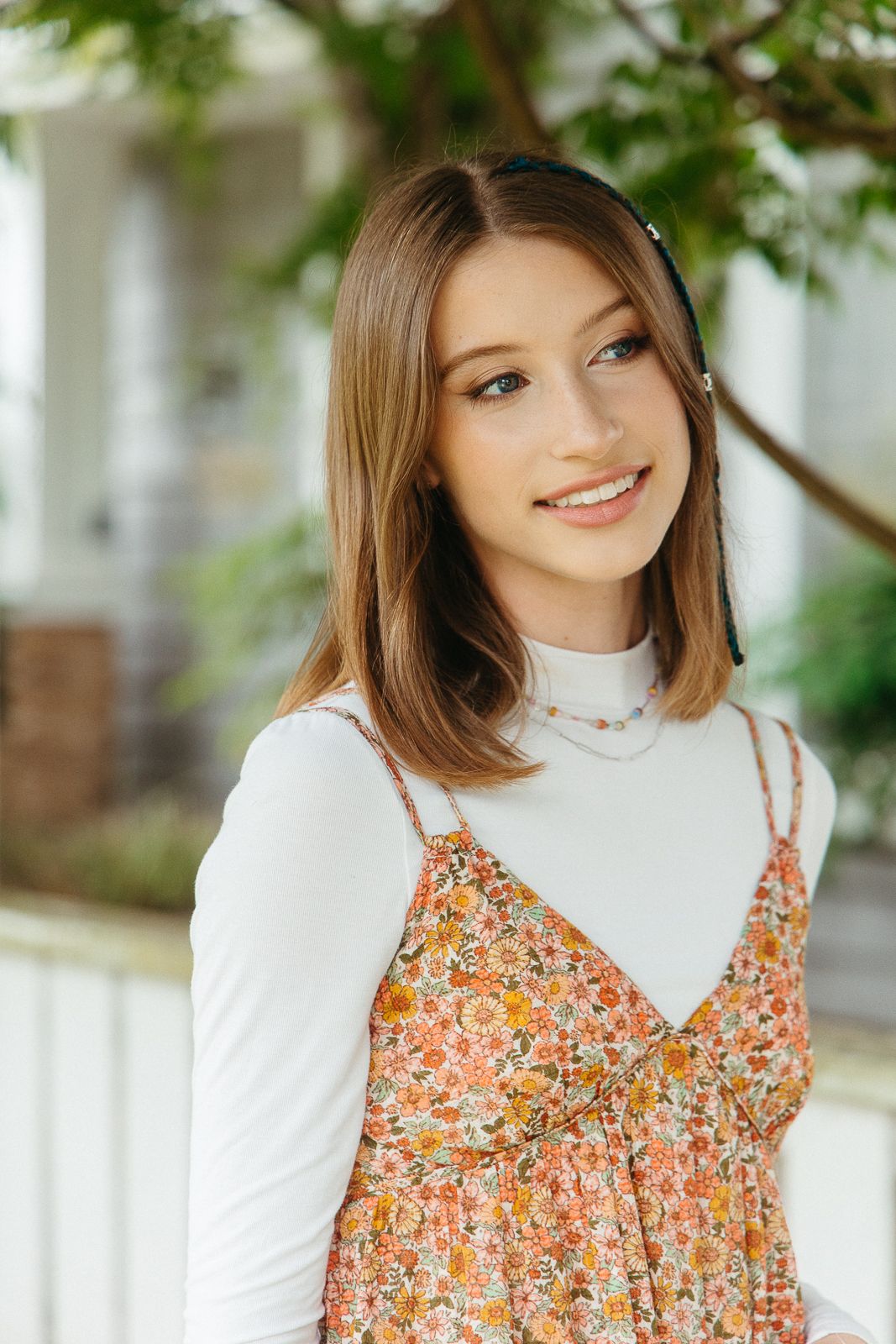 Young woman in white turtleneck and floral dress standing near greenery.
