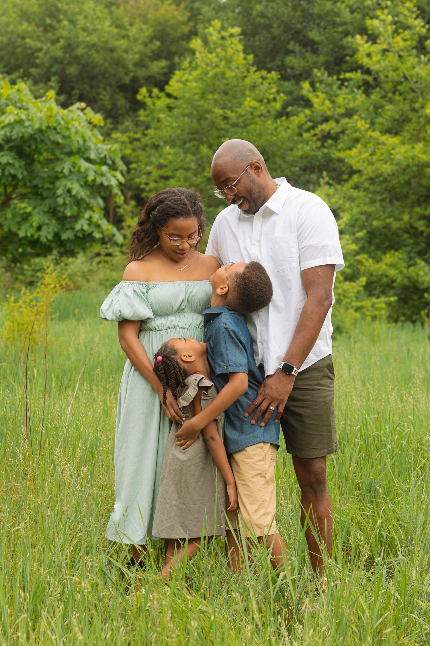 A family shares a tender moment together while standing in a sunlit meadow filled with tall grass and greenery.