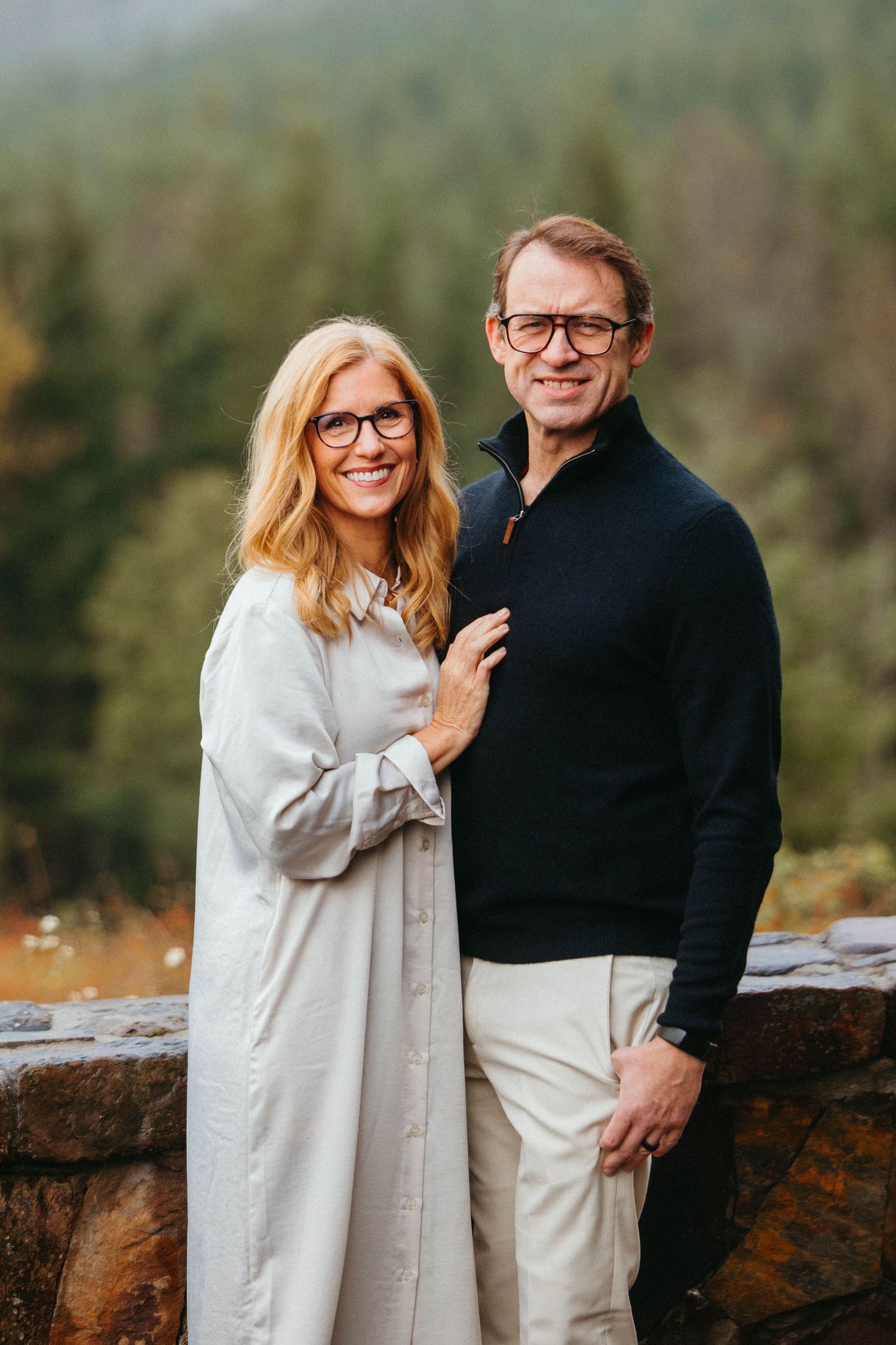 A couple in casual attire standing together on an outdoor viewpoint with forest backdrop.