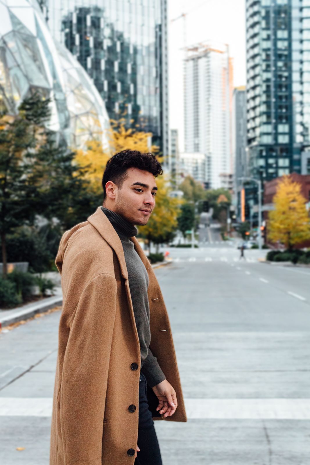 Street fashion photo with camel coat against modern city architecture and fall foliage.