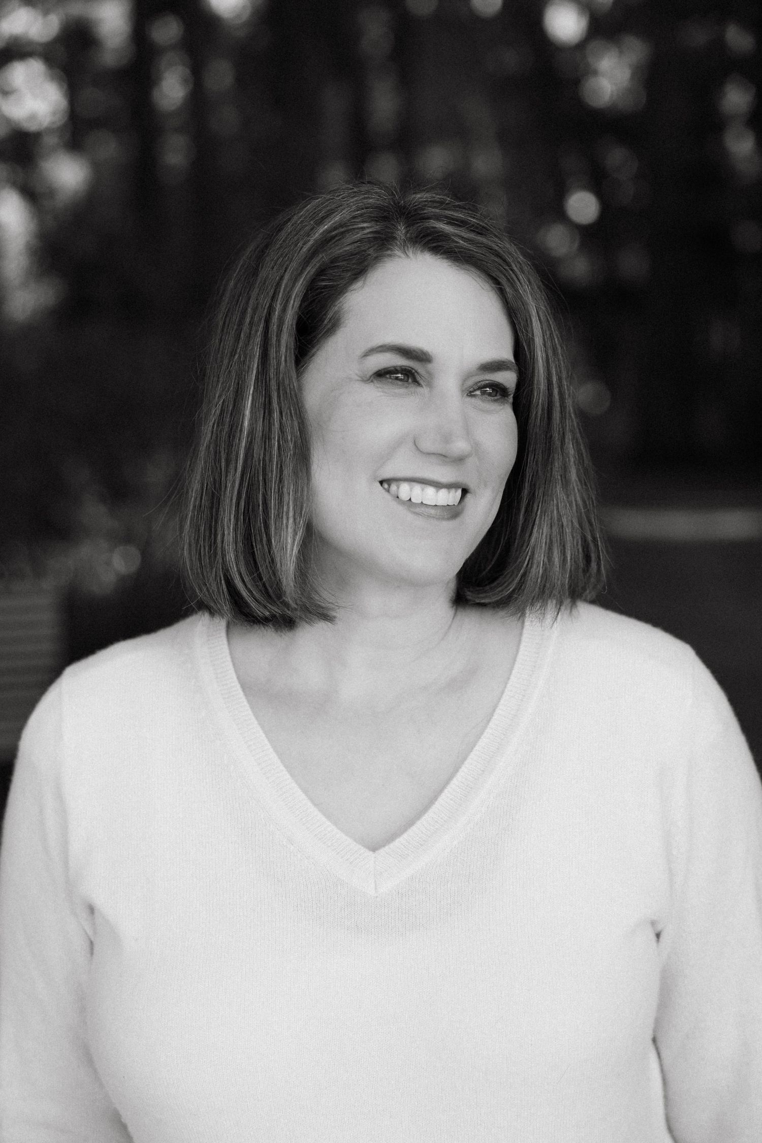 Black and white portrait of a smiling person with shoulder-length hair wearing a white v-neck sweater.