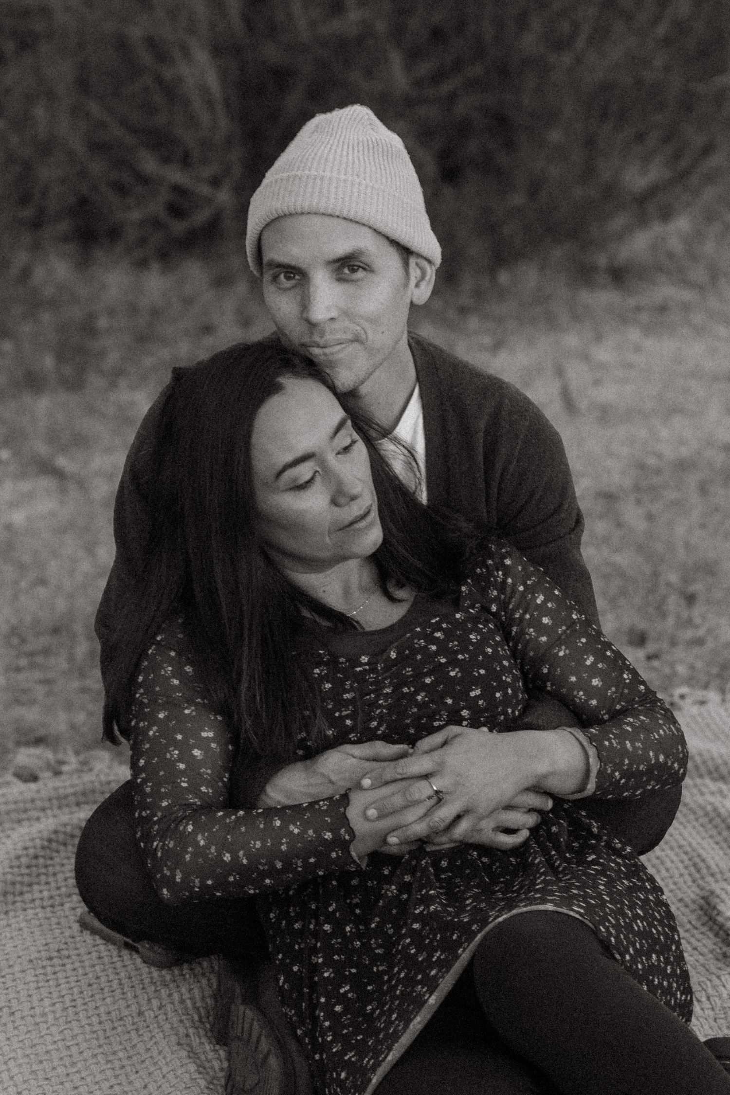 A black and white photo of a couple cuddling on a picnic blanket outdoors.
