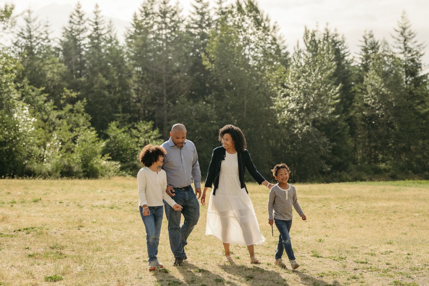 Family walking together and holding hands in a sunny field with evergreen trees in the background on a summer day.