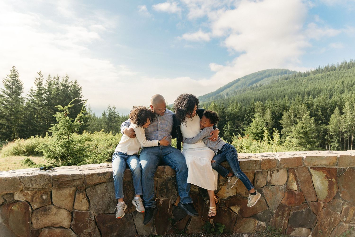 A group sits together on a stone wall overlooking a scenic mountain landscape with evergreen forests on a sunny day.