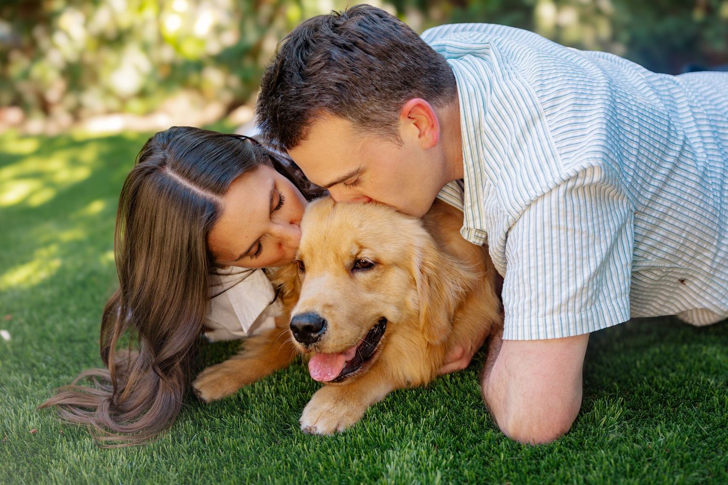 Couple cuddling with golden retriever dog on sunny lawn.
