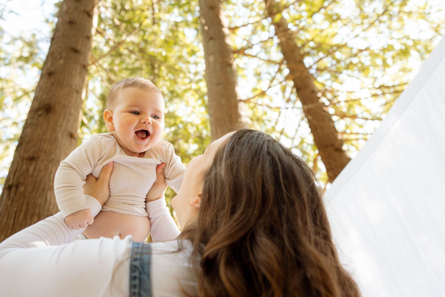 A heartwarming moment between parent and happy baby beneath tall trees on a bright sunny day.