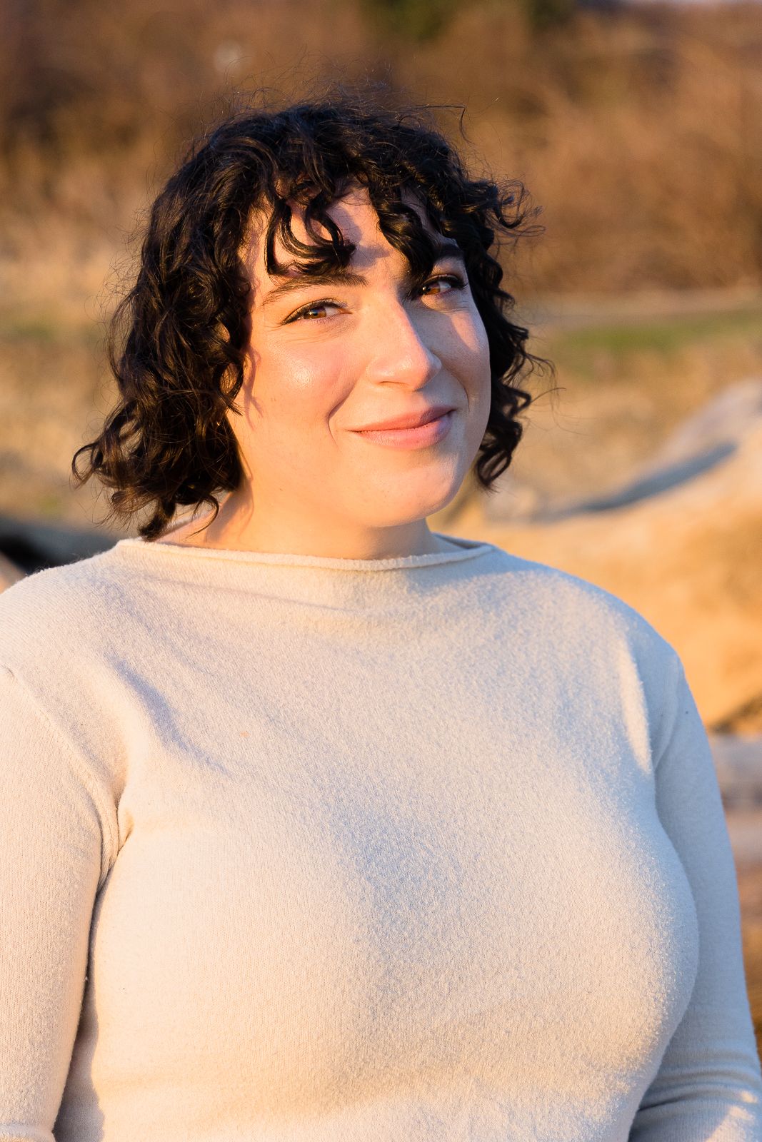 Woman in white sweater smiling warmly in golden outdoor lighting.