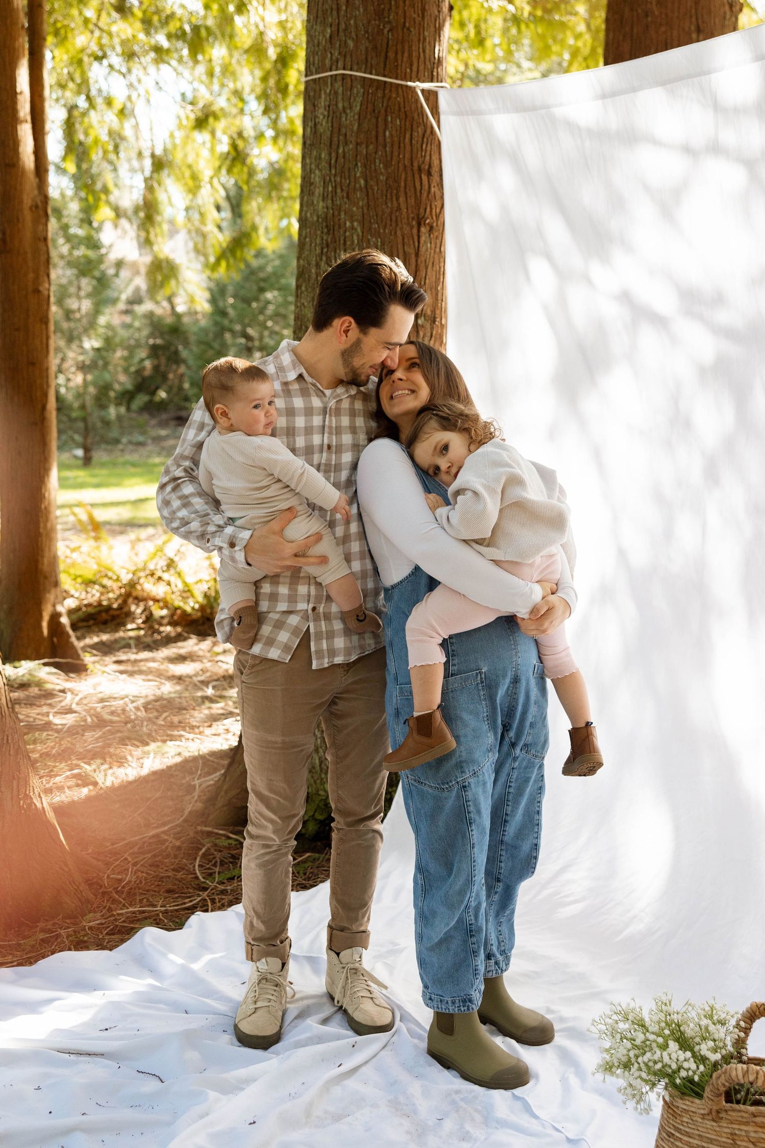 A family embraces tenderly in front of a white backdrop outdoors, surrounded by sunlit trees and nature.
