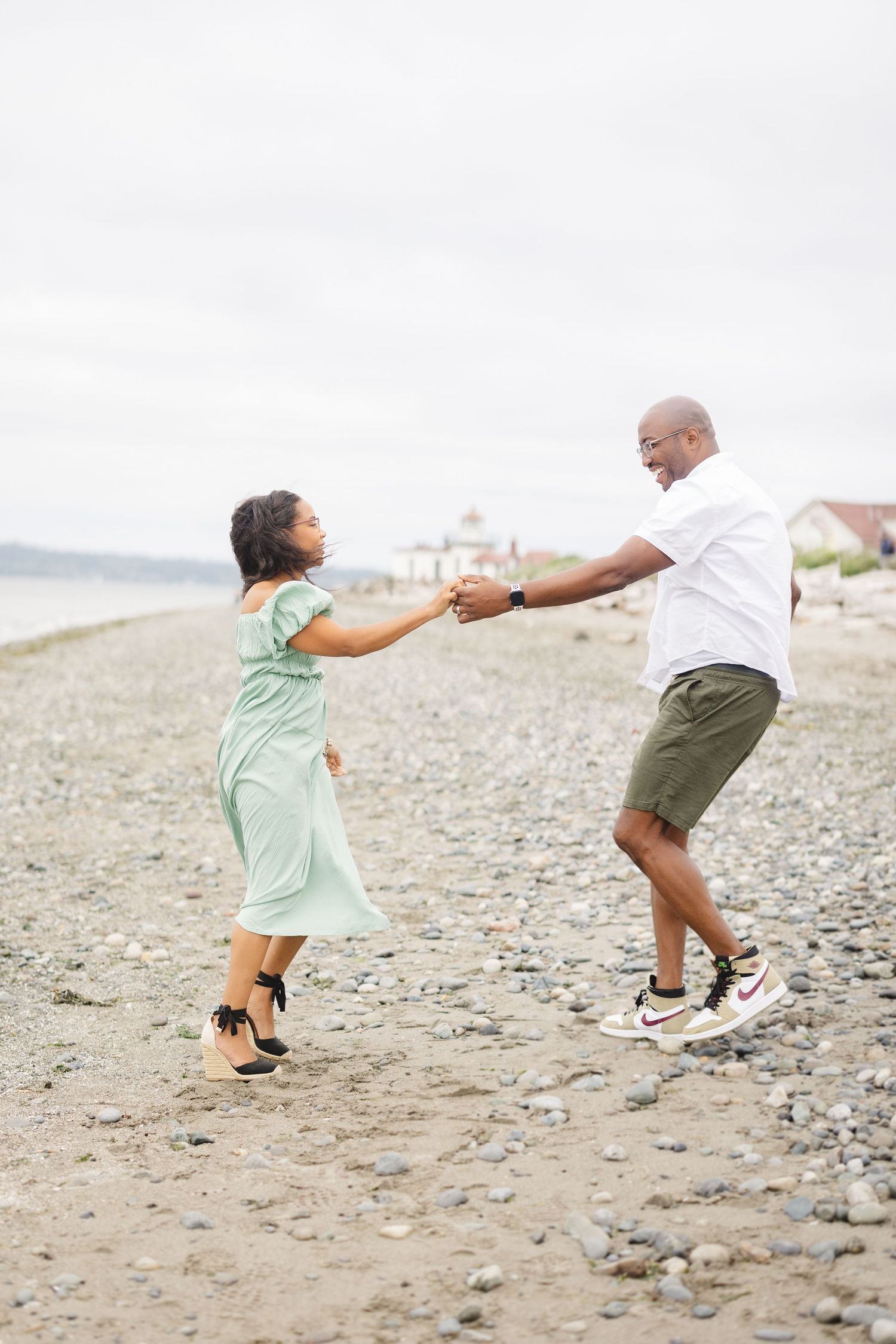 Playful couple dancing and holding hands on rocky beach with cloudy sky.