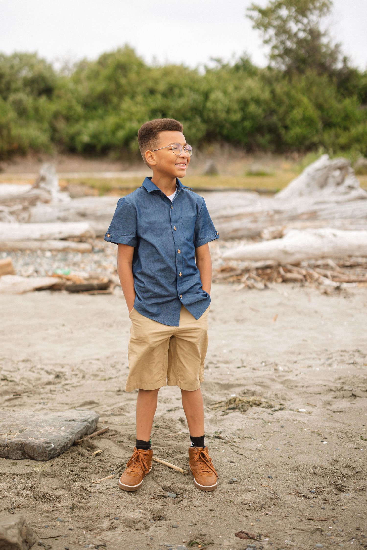 Young boy in blue shirt and khaki shorts stands on sandy beach with driftwood and green foliage in background.