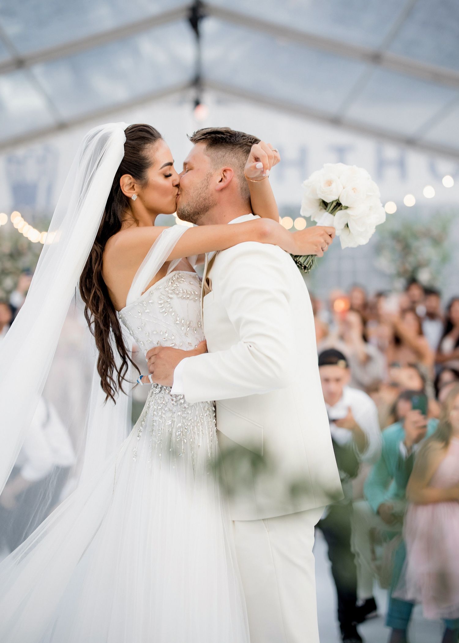 Bride and groom share first kiss at elegant outdoor wedding under glass tent with string lights and white floral bouquet.