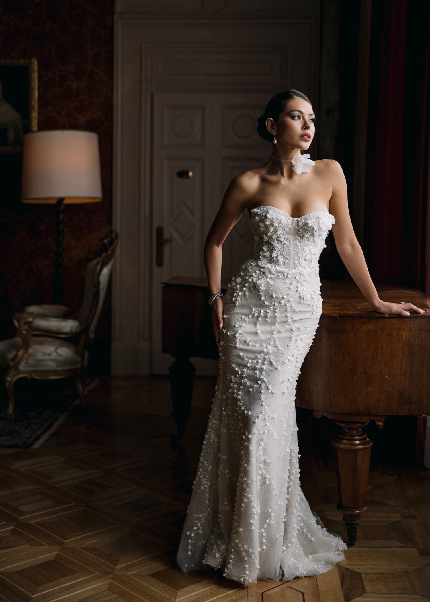 Bride in strapless floral appliqué white mermaid wedding gown posing elegantly in a dimly lit vintage room.