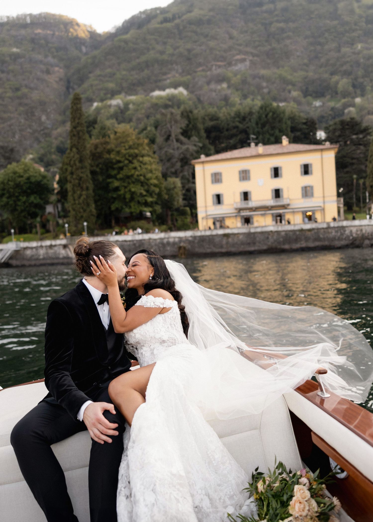Bride and groom kissing on a wooden boat on Lake Como, Italy, with lush green hills and a yellow villa in the background.